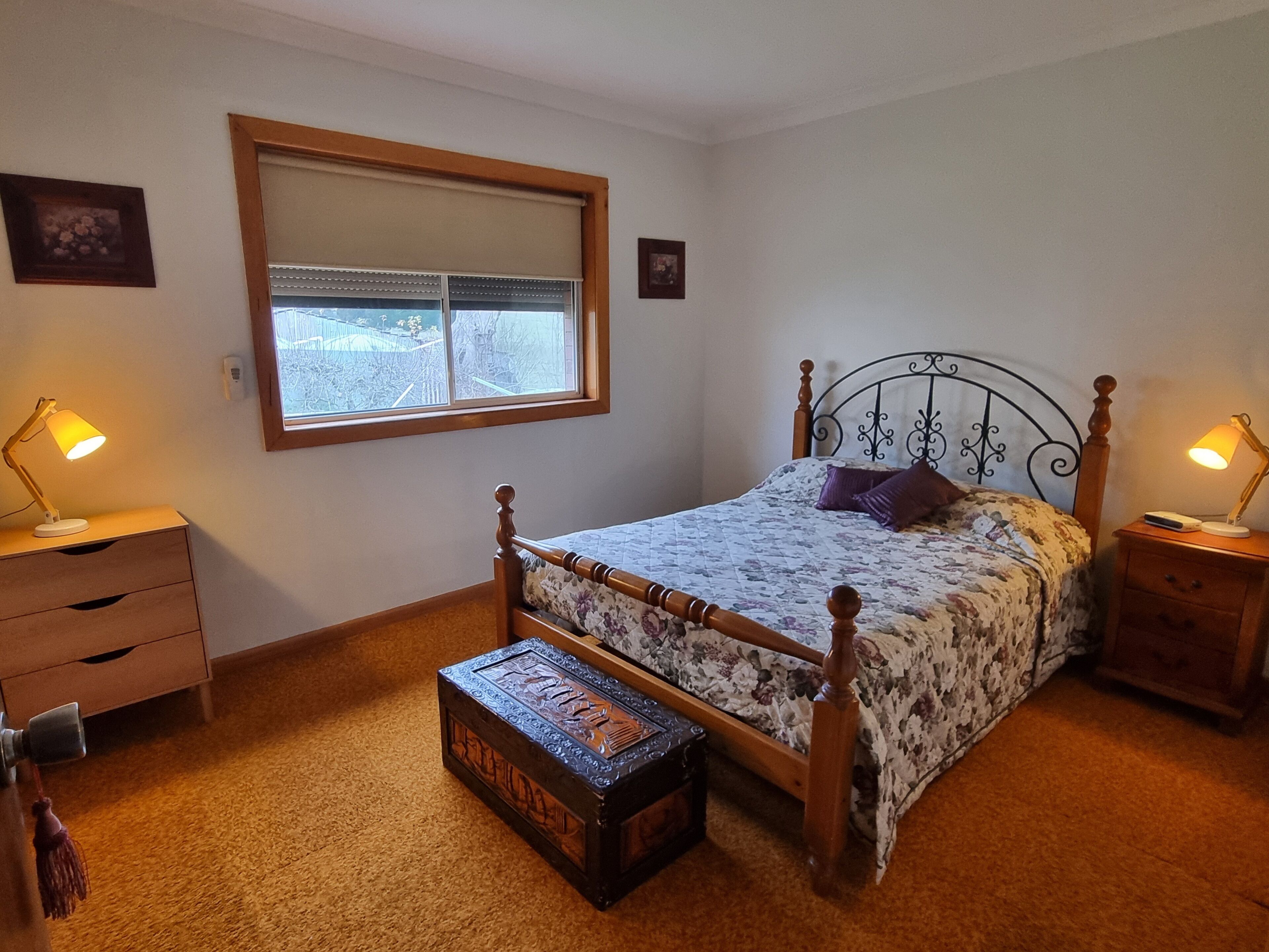 Un dormitorio con una cama con dosel, un cofre de madera, dos mesitas de noche con lámparas, una ventana con persianas y un cuadro pequeño en la pared.