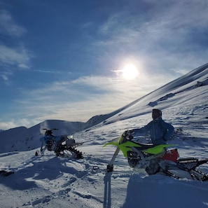 Une montagne enneigée, un soleil éclatant dans le ciel et une personne sur une moto verte.