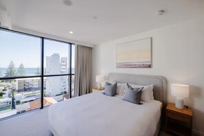 A hotel room with a large bed, two bedside tables with lamps, a painting on the wall, and a view of buildings and trees through the window.