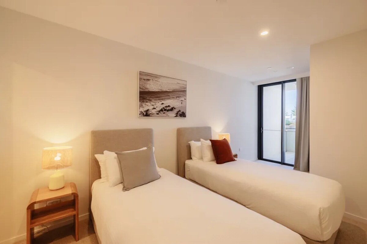Ein Hotelzimmer mit zwei Betten, einem Nachttisch mit Lampe, einem Bild an der Wand und einem Fenster mit Vorhängen.