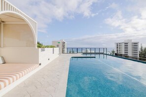 Ein Pool auf dem Dach mit freiem Blick auf das Meer, eine überdachte Sitzgelegenheit und moderne Gebäude im Hintergrund.
