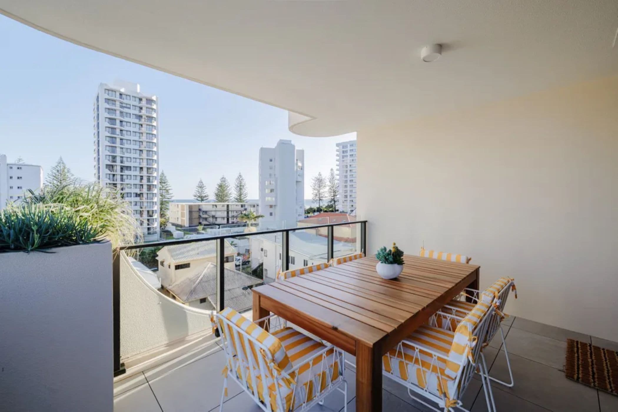 Un balcón con mesa y sillas de madera, una planta en maceta y vistas de edificios circundantes.