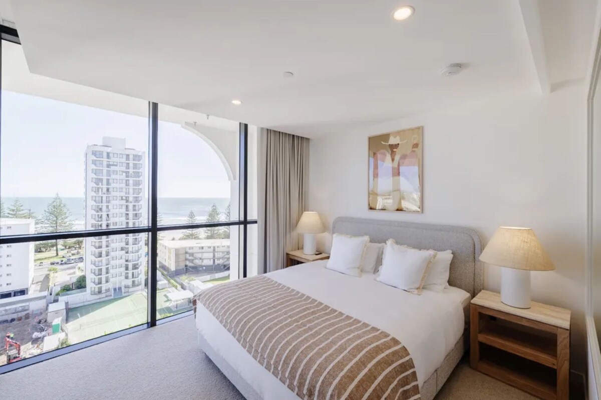 Ein Hotelzimmer mit einem großen Bett, zwei Nachttischen mit Lampen, einem Bild an der Wand und Blick auf die Stadt durch raumhohe Fenster.