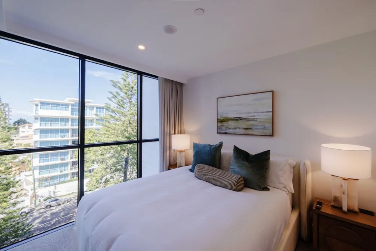 Ein Hotelzimmer mit einem großen Bett, zwei Nachttischlampen, einem Bild an der Wand und Blick auf Gebäude und Bäume durch das Fenster.