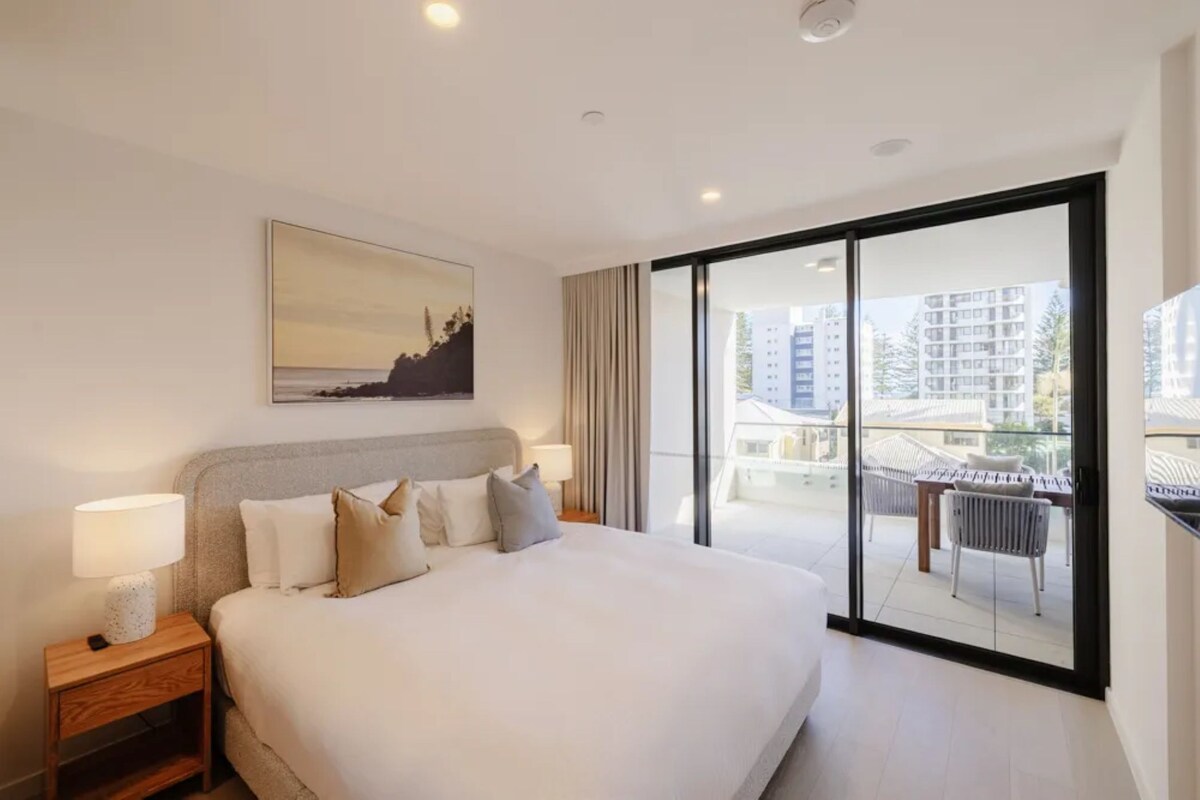 Ein Hotelzimmer mit einem großen Bett, einem Nachttisch mit Lampe, einem Bild an der Wand und Blick auf die Stadt durch eine Glastür.