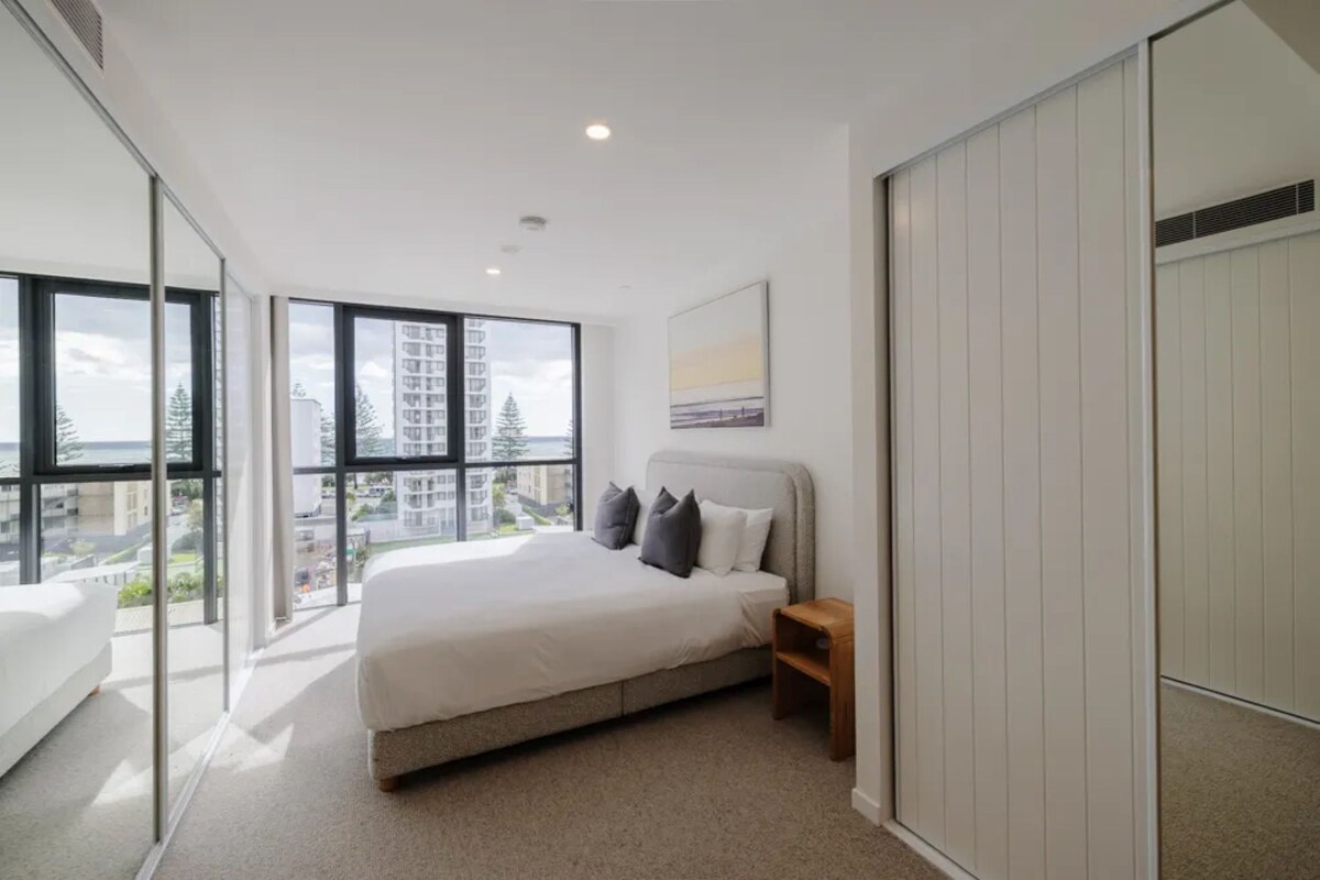 Ein modernes Schlafzimmer mit einem großen Bett, einem Nachttisch, einem Kleiderschrank und Blick auf die Stadt durch raumhohe Fenster.