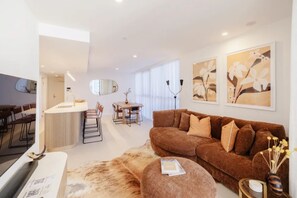 A modern living room with a brown sofa, a dining area with a table and chairs, and a kitchen area with a sink and counter.