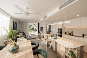 A modern kitchen with a central island, a dining area with a wooden table and chairs, and a living space with a sofa and a ceiling fan.