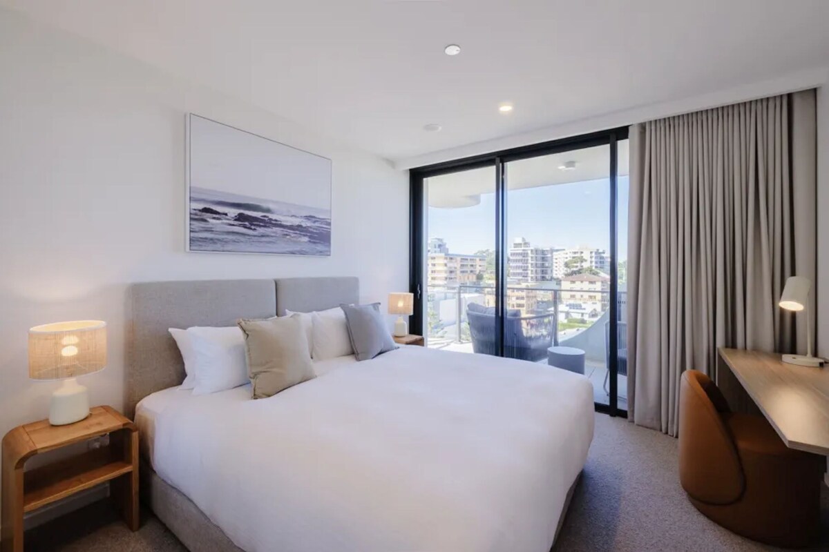 Ein Hotelzimmer mit einem großen Bett, einem Schreibtisch, einem Sessel, einem Nachttisch, einer Lampe, einem Fenster mit Blick auf Gebäude und einer Deckenleuchte.