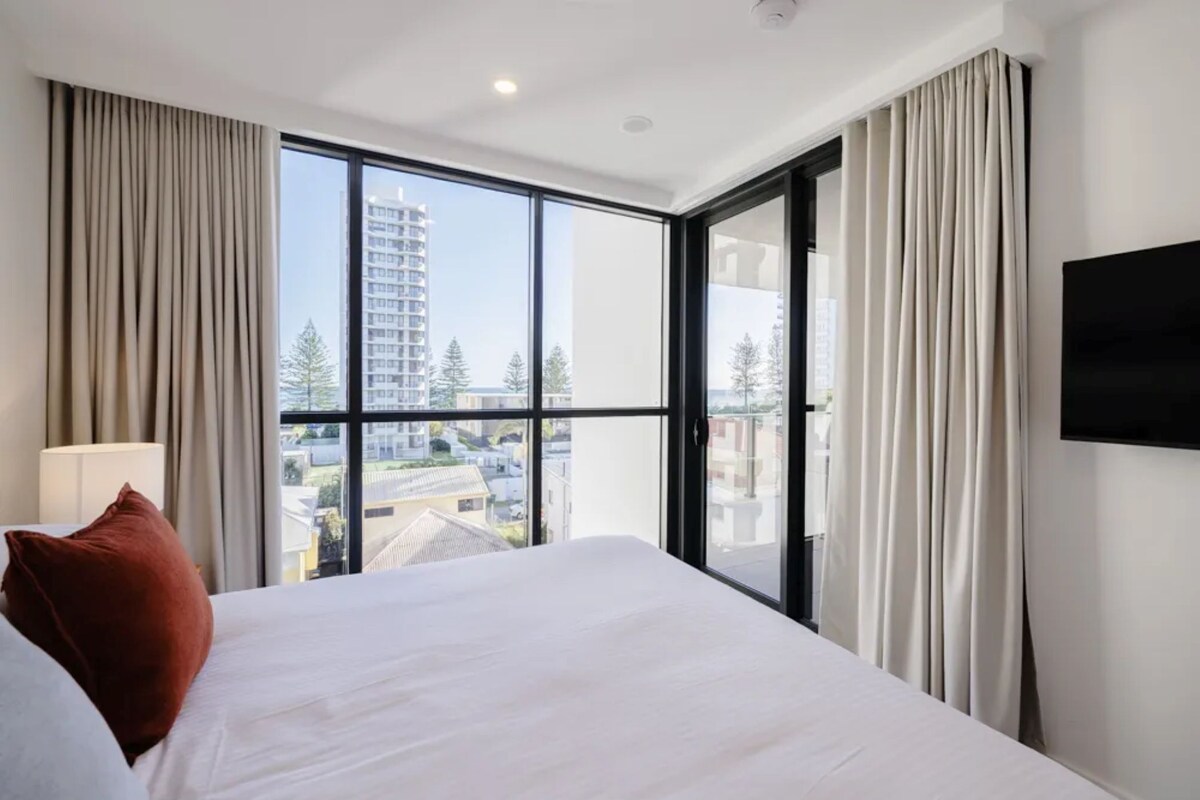 Ein Hotelzimmer mit einem großen Bett, einem Flachbildfernseher und Blick auf Gebäude und Bäume durch raumhohe Fenster.