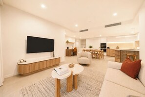 Ein modernes Wohnzimmer mit Flachbildfernseher, einer Couch, einem Couchtisch und einem Essbereich im Hintergrund.