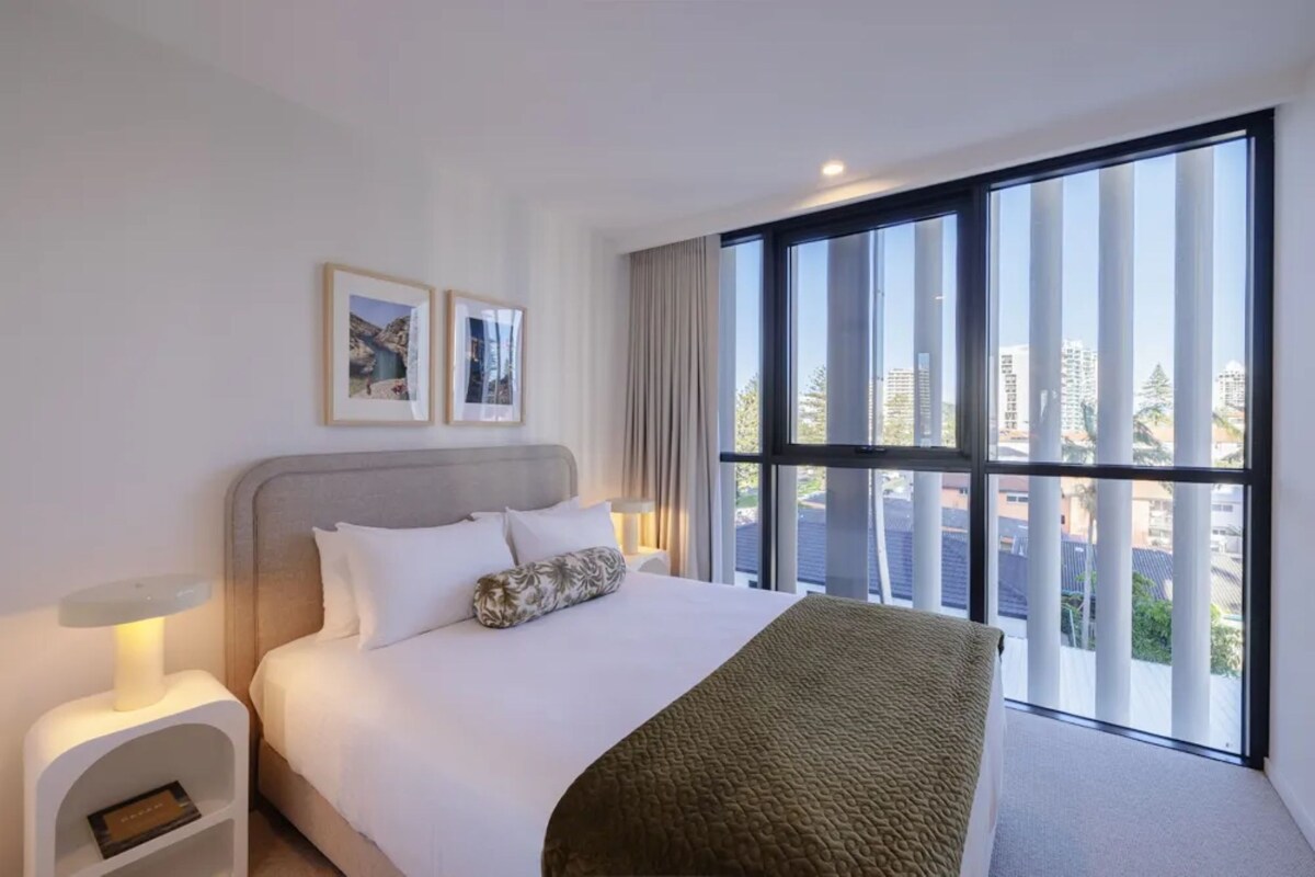 Ein Hotelzimmer mit einem großen Bett, einem Nachttisch, einem Fenster mit Blick auf die Stadt und zwei gerahmten Bildern an der Wand.