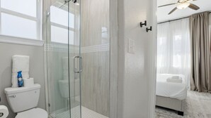 Un baño de hotel con ducha de vidrio, inodoro y cama con colcha blanca.