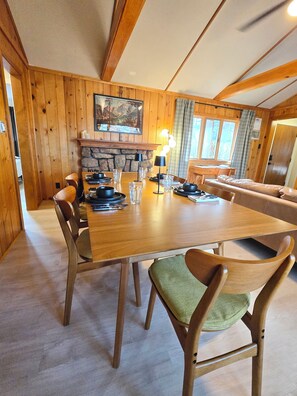 A wooden dining area with a table set for four, chairs, a stone fireplace, and a painting on the wall.