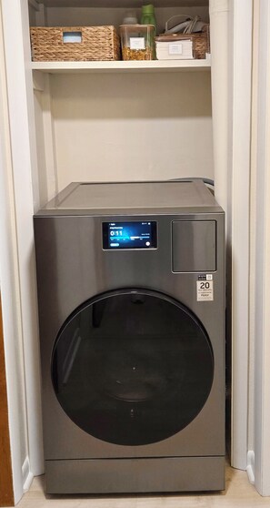 A compact, stainless steel front-loading washing machine with a digital display, situated in a laundry room with a white door and a shelf above containing baskets and containers.