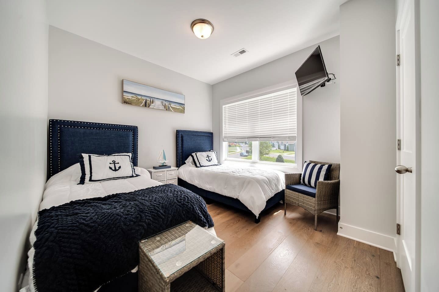 A bedroom with two beds, a television, a window with blinds, and a wicker chair.
