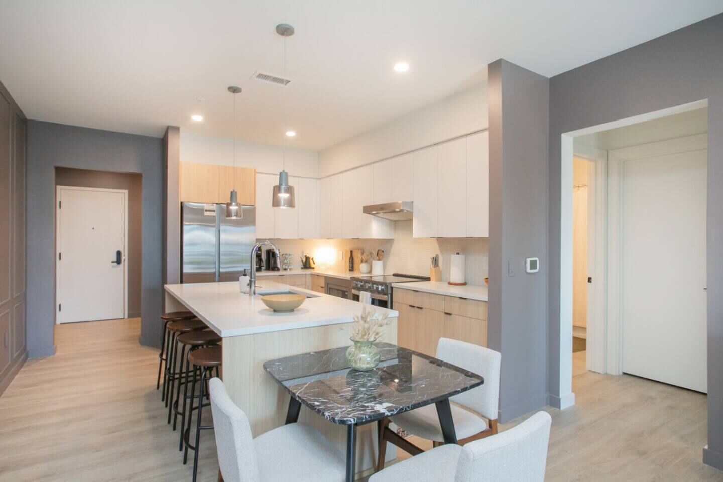 A modern kitchen with a central island, bar stools, a dining table with chairs, and a view into the adjacent living area.