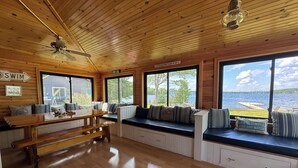 Una cabaña con paredes y techo de madera, ventanas amplias con vistas a un lago, una zona de comedor con banco y un ventilador de techo.