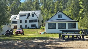 Una casa blanca de dos pisos con techo de metal, un edificio más pequeño adosado, una mesa de picnic y un vehículo rojo estacionado al frente.