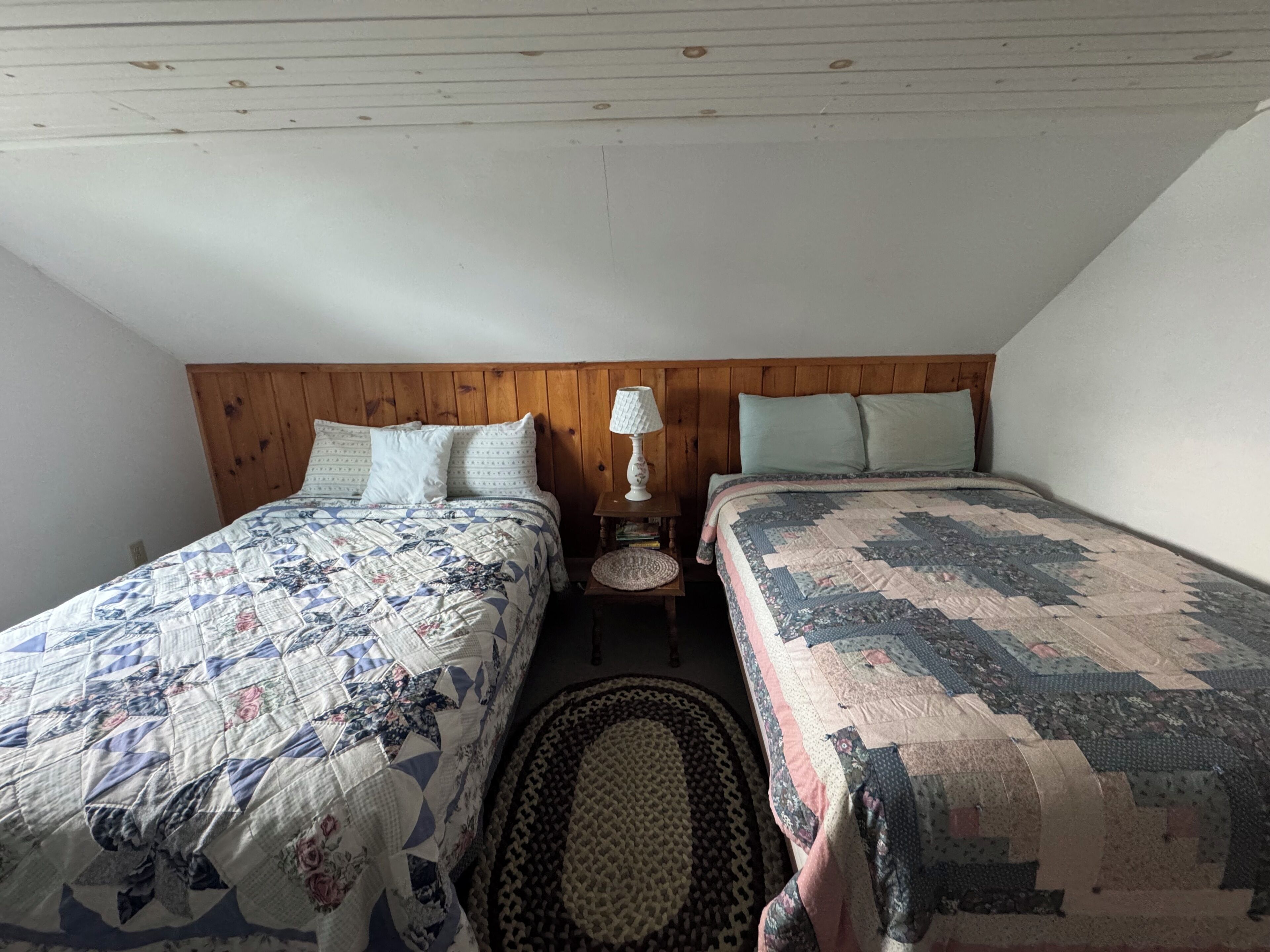 A bedroom with two beds, a wooden headboard, a bedside table with a lamp, and a patterned rug.