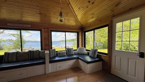 Una cabaña con interior de madera, grandes ventanales con vista a un lago, bancos integrados con cojines y una puerta con ventana.