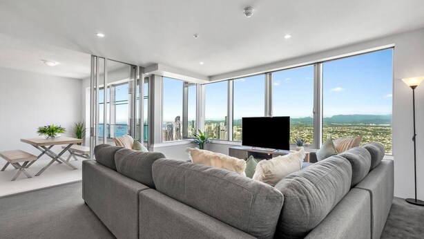 A modern living room with a large gray sectional sofa, a flat-screen TV, and a dining area with a table and chairs. - Elevé at Q1 - Spectacular Beach + Hinterland views (Surfers Paradise)