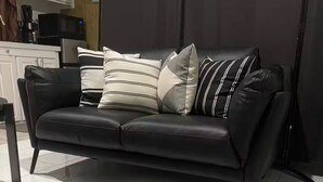 En svart lädersoffa med randiga kuddar, en vävd matta och ett köksområde i bakgrunden.