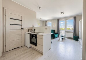 Eine moderne Küche mit integriertem Backofen und einer kleinen Kochinsel, ein Essbereich mit einem grünen Sofa und einem Tisch, und ein großes Fenster mit Blick auf den Himmel und das Grün.