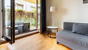 Ein modernes Zimmer mit grauem Sofa, einem kleinen runden Tisch, einer Stehlampe und einer Glastür, die zu einem Außenbereich führt.