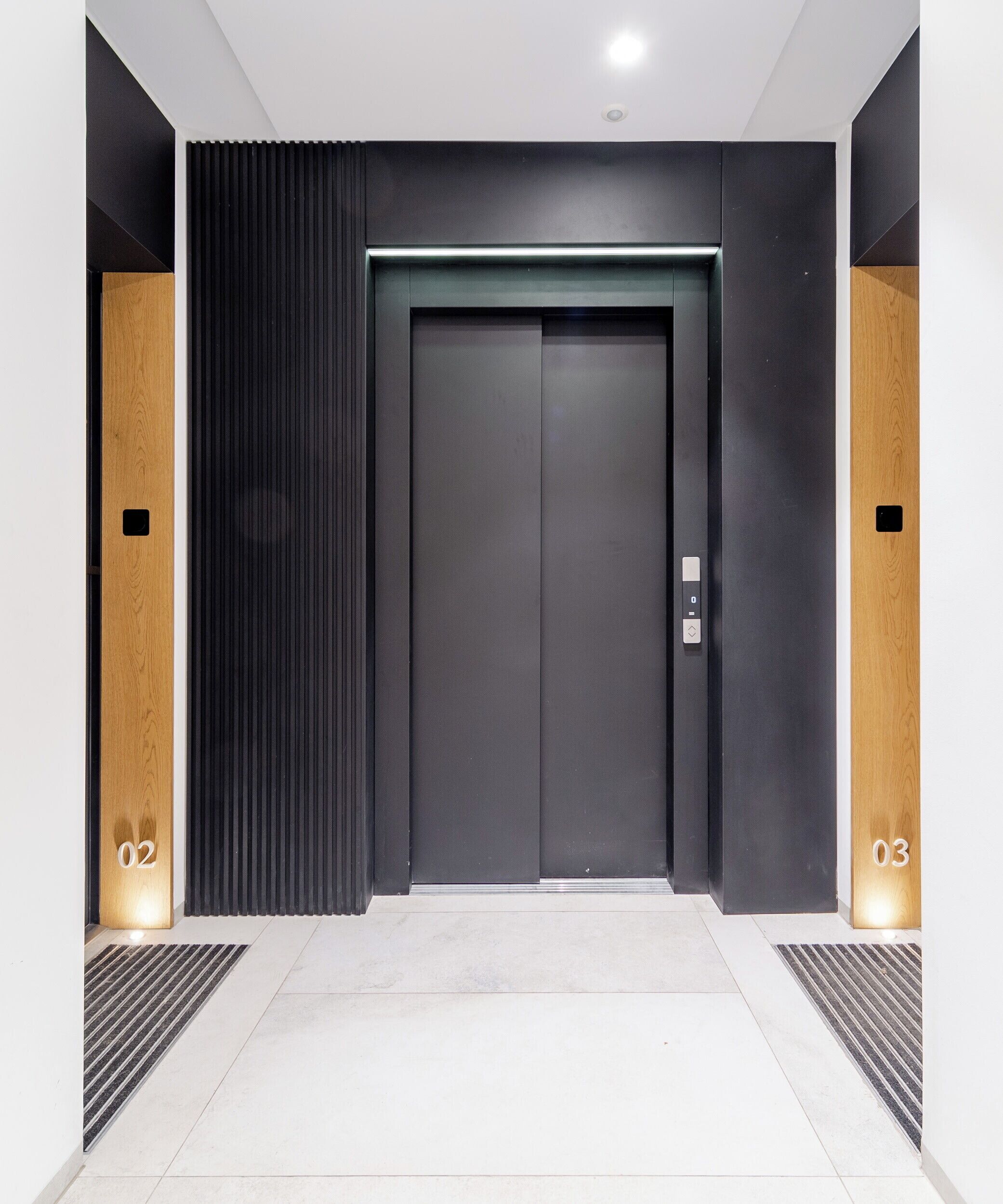 A modern elevator with a dark door and a control panel, flanked by vertical wooden panels and a light-colored floor.
