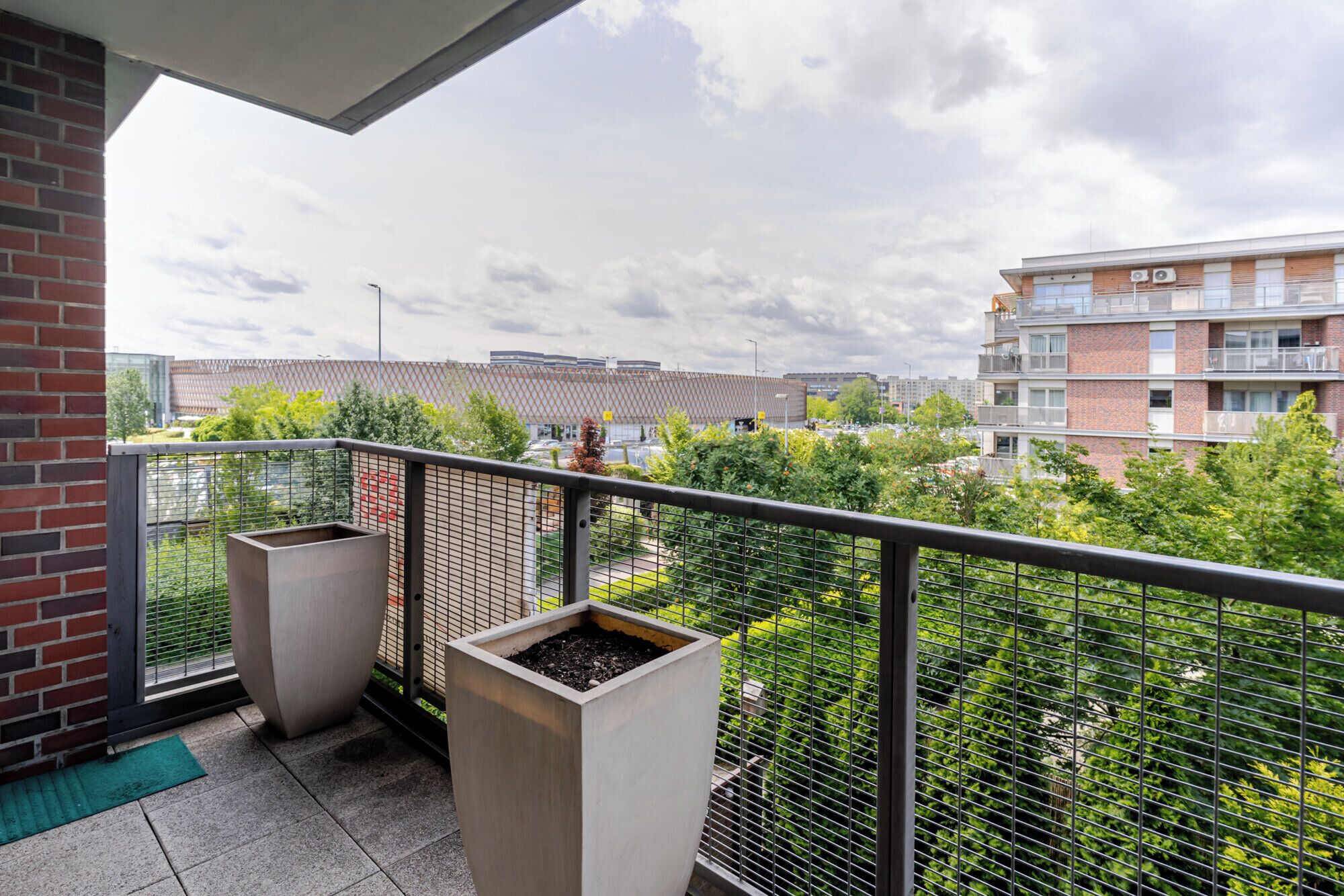 Een balkon met twee plantenbakken, een metalen hek en uitzicht op een woongebouw en bomen.