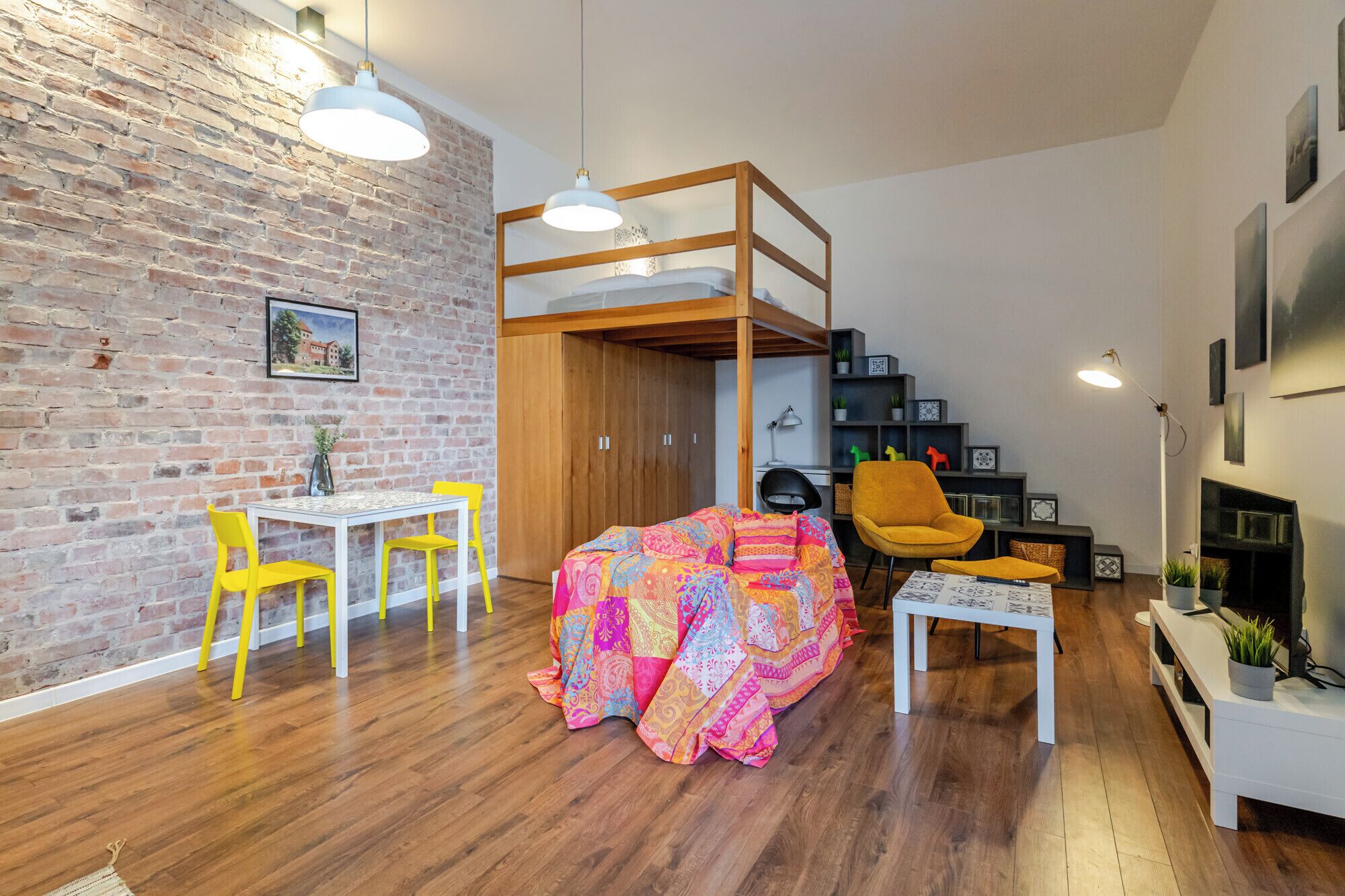 Una stanza con parete in mattoni, pavimento in legno, un letto a castello, una scrivania, una sedia e un tavolino con una coperta colorata.
