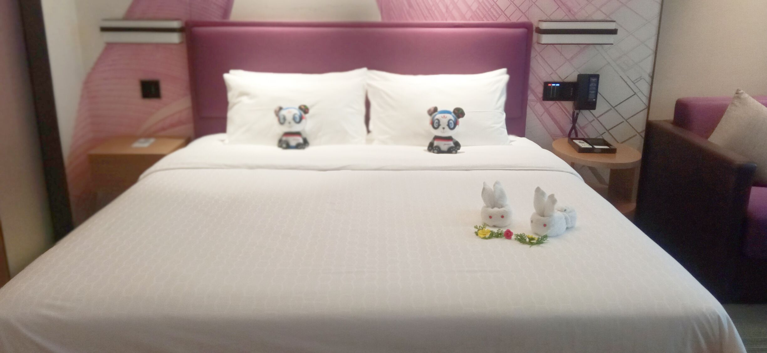 Una camera d'albergo con un letto, due cuscini decorati con motivi panda, un comodino con un telefono e una sedia.