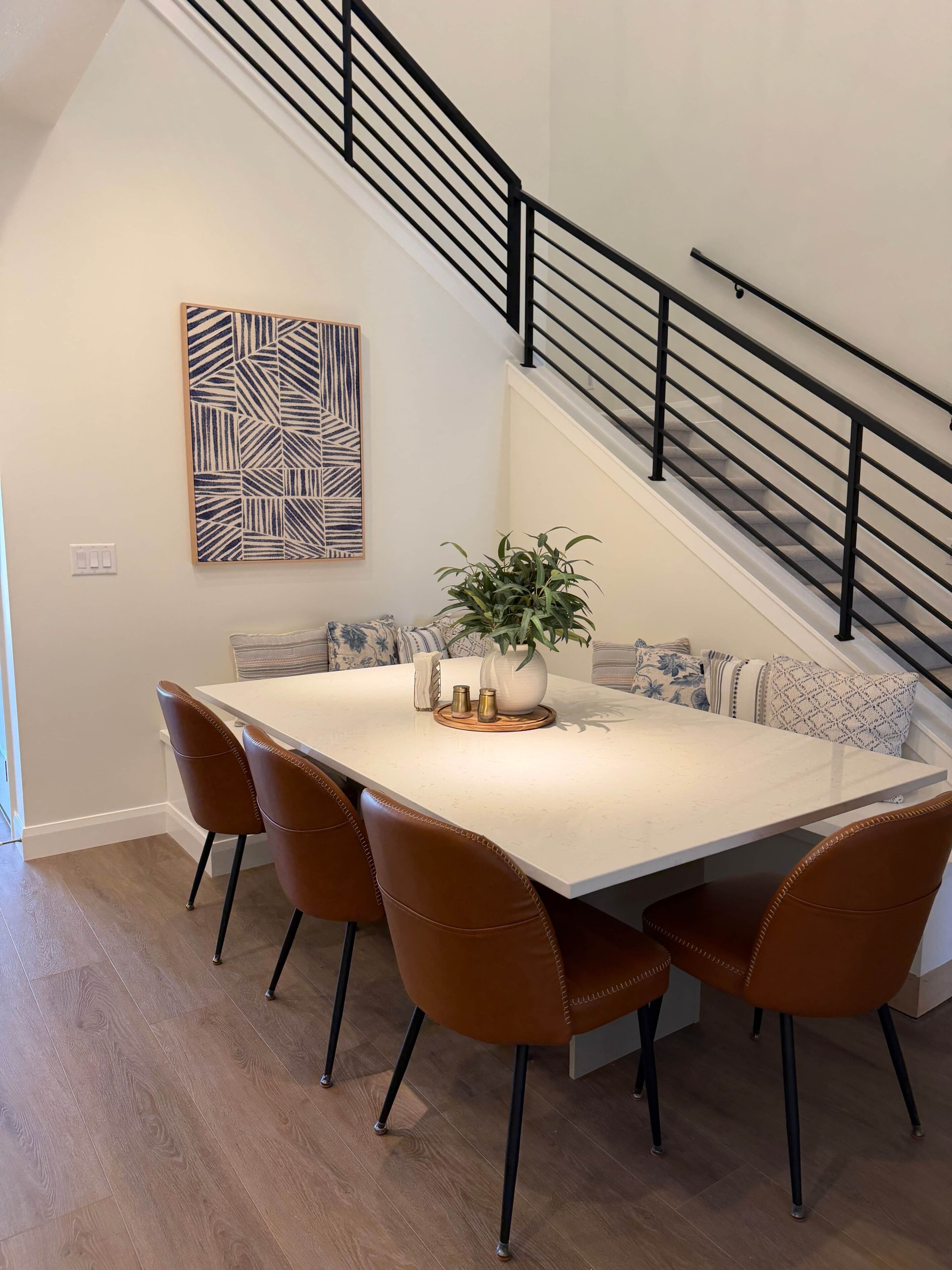 A dining area with a white table, brown chairs, a plant centerpiece, and a staircase with a black railing.