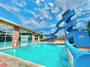 A swimming pool with a water slide, a brick building with large windows, and a clear blue sky. - Holiday Park Home Sleeps 4 - Dogs Welcome - Pool (Clacton-on-Sea)