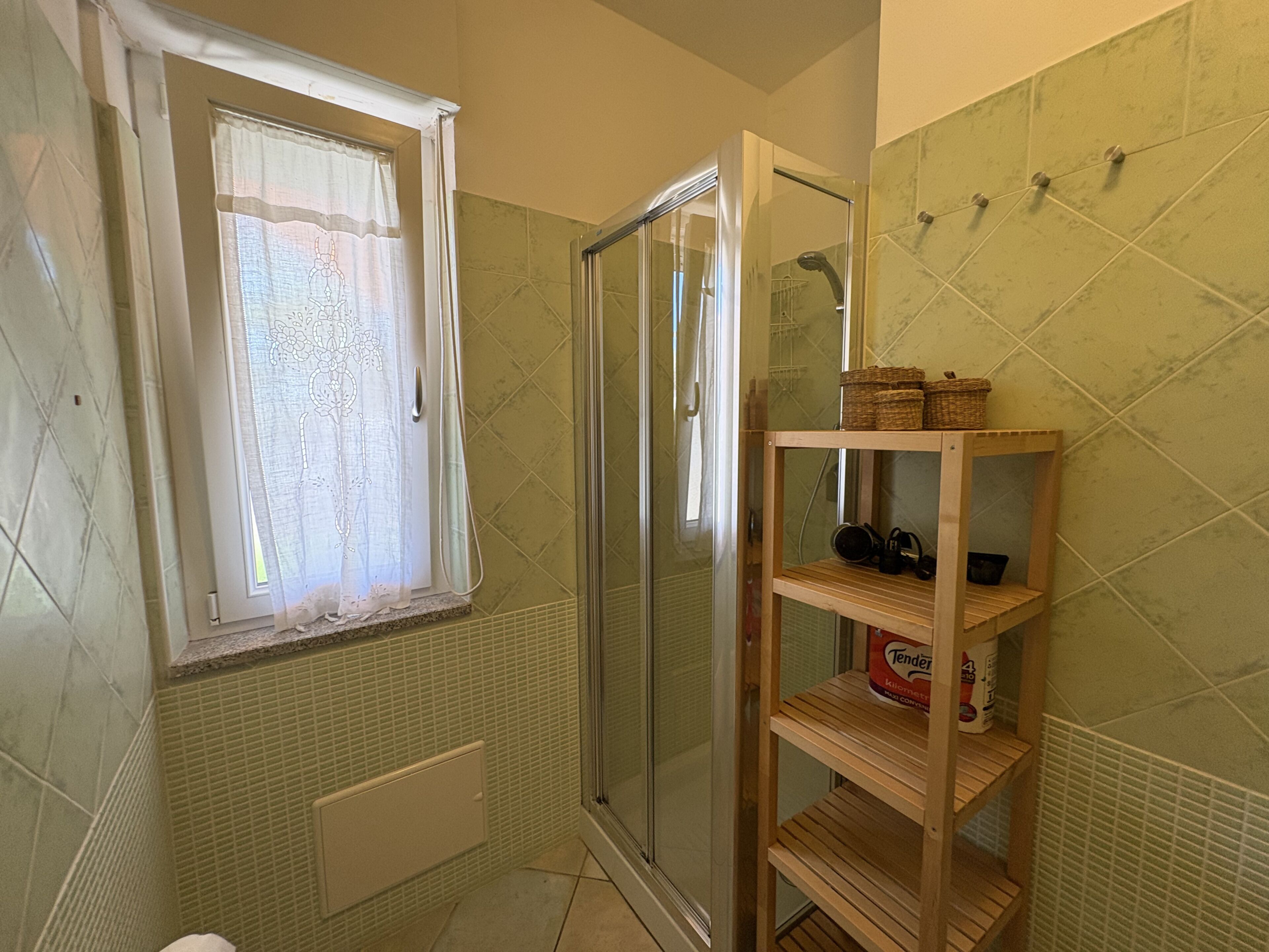 Ein Badezimmer mit Dusche, einem Fenster mit Vorhang und einem Holzregal mit Körben.