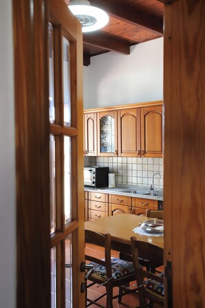 A wooden-framed door leading into a kitchen with wooden cabinets, a tiled backsplash, a stove, a microwave, and a dining table with chairs. - Sa Domu De Cadeddu - Three Bedroom Apartment, Sleeps 5 (Nurachi)