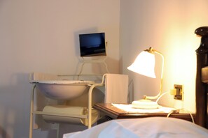 Ein Krankenzimmer mit einem Bett, einem Nachttisch mit Lampe, einem Monitor an der Wand und einem Waschbecken.