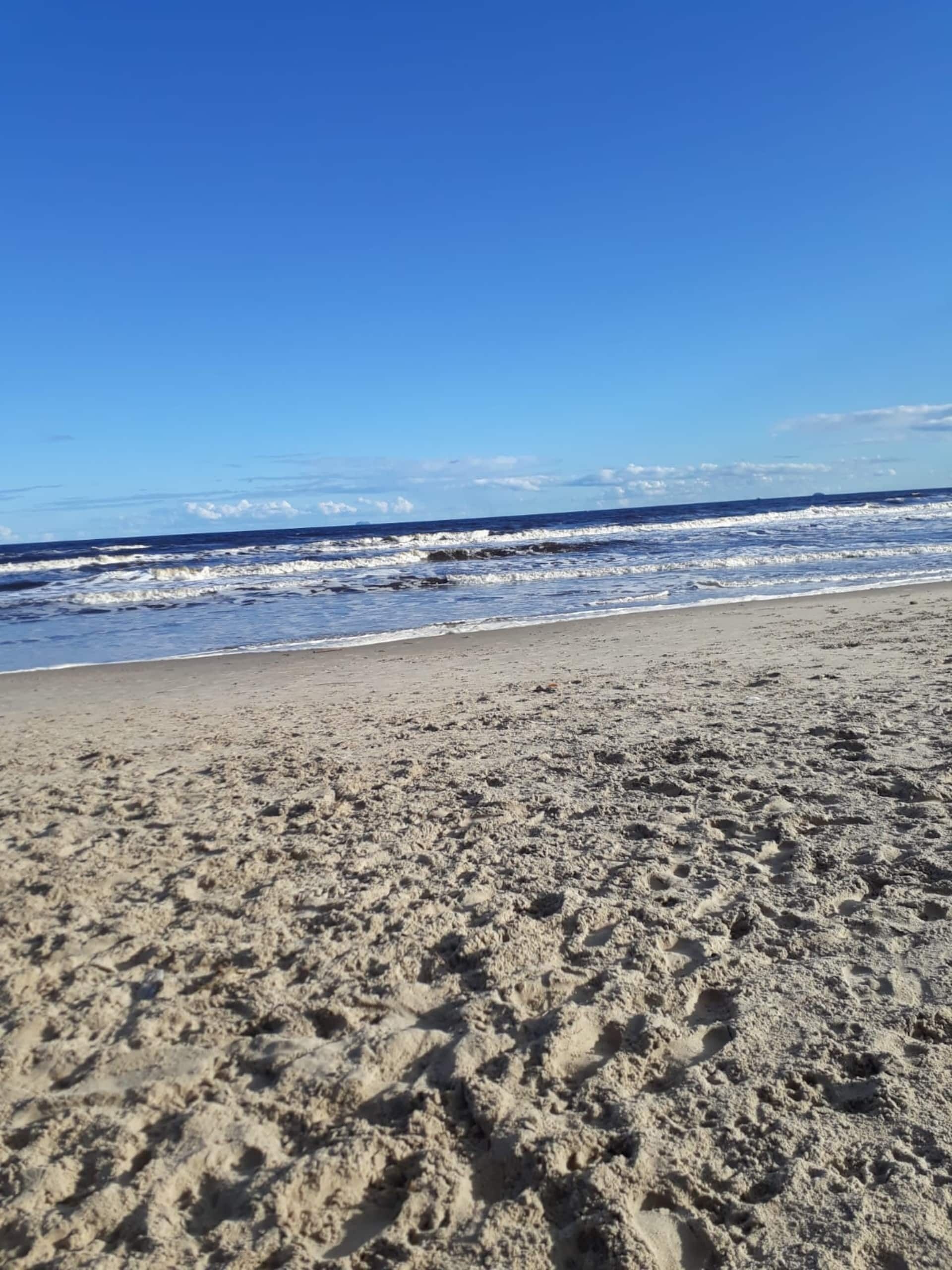 Uma praia arenosa com pegadas que levam à água, sob um céu azul e límpido.