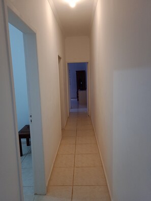 A narrow hallway with white walls and a tiled floor, leading to a dark room at the end.
