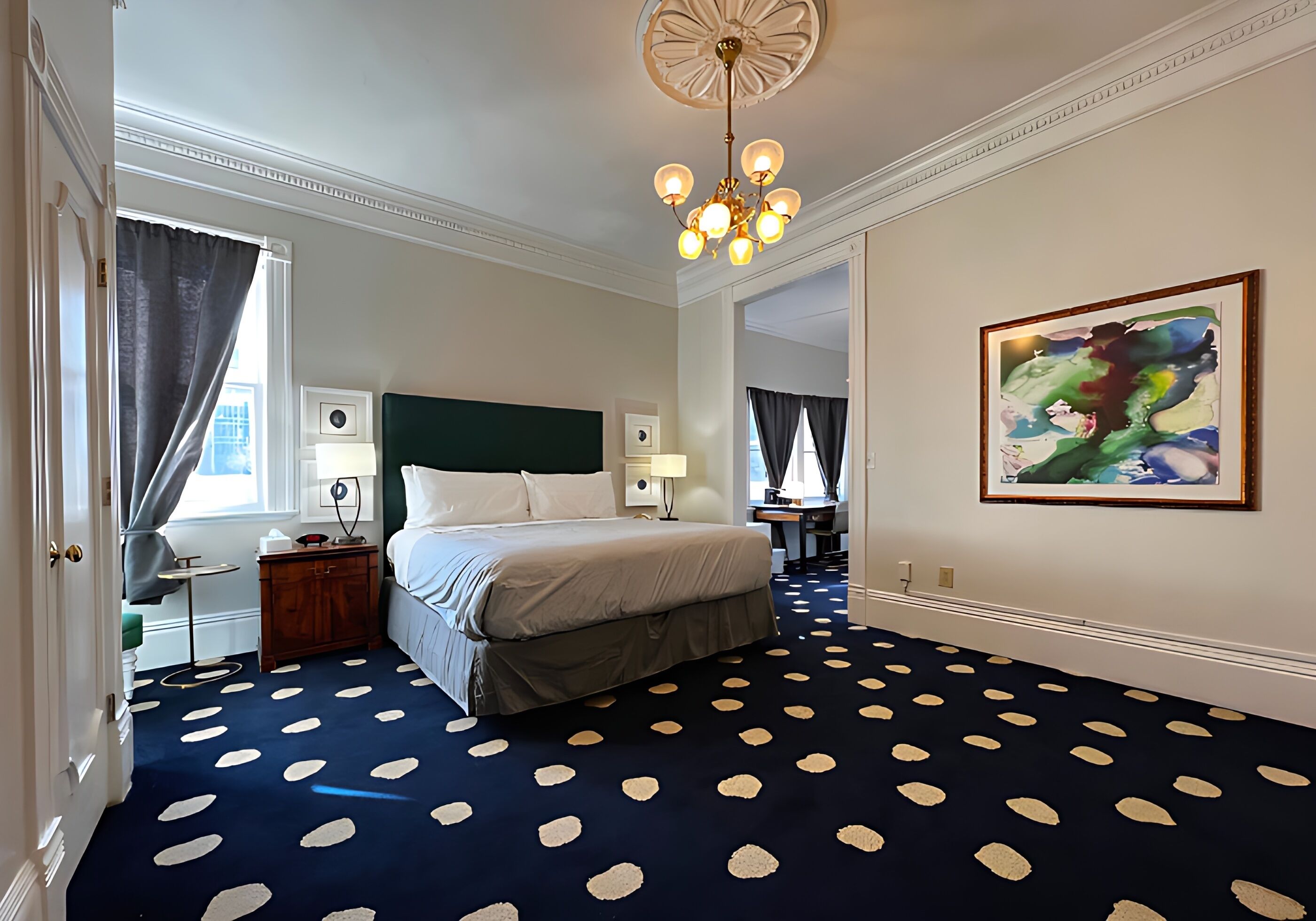 Habitación de hotel con cama, escritorio, silla, lámpara, un cuadro y alfombra estampada.