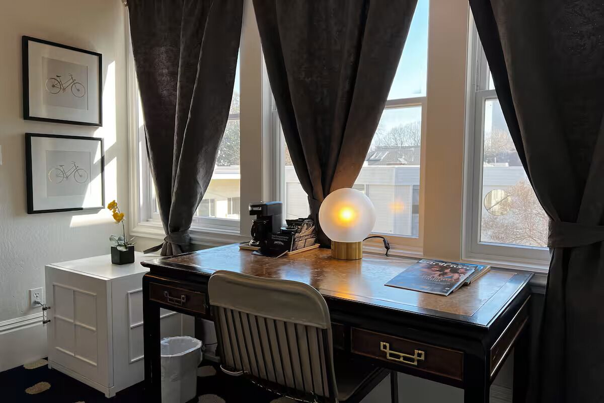 Una habitación con escritorio, silla, ventana con cortinas, cuadros enmarcados y una lámpara.