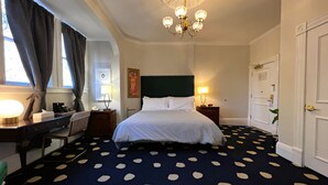 Habitación de hotel con cama, escritorio, silla, lámpara de escritorio, reloj de pared, cuadro, candelabro y puerta.