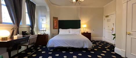 Quarto de hotel com cama, escrivaninha, cadeira, luminária de mesa, relógio na parede, pintura, lustre e porta.