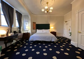 Habitación de hotel con cama, escritorio, silla, lámpara de escritorio, reloj de pared, cuadro, candelabro y puerta.