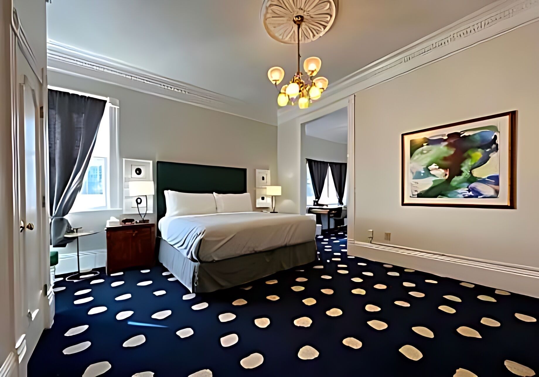 Habitación de hotel con cama, escritorio, silla, lámpara de escritorio, obra de arte colgada en la pared y alfombra estampada.