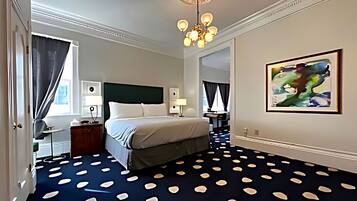 Habitación de hotel con cama, escritorio, silla, lámpara de escritorio, obra de arte colgada en la pared y alfombra estampada.