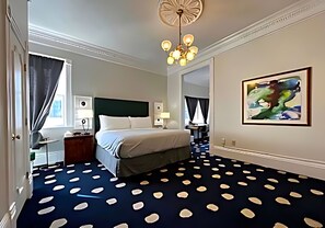 Habitación de hotel con cama, escritorio, silla, lámpara de escritorio, obra de arte colgada en la pared y alfombra estampada.
