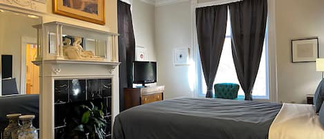 Una habitación de hotel con cama, televisión, chimenea, espejo y ventana con cortinas.
