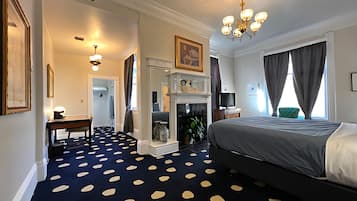 Habitación de hotel con cama, chimenea, escritorio y un amplio ventanal con cortinas.